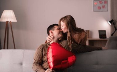 Evdeki kanepede öpüşen genç çift. Kadın ona arkadan sürpriz yaparken adam kalp şeklinde balon tutuyor. Aşk ve aşk kavramı. Evde sevgililer gününü kutluyoruz..