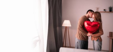 Genç, mutlu bir çift birbirlerine sarılıyor ve öpüşüyorlar. Evde kalp şeklinde balon tutuyorlar. Neşeli bir Sevgililer Günü konsepti. Pankart. Boşluğu kopyala