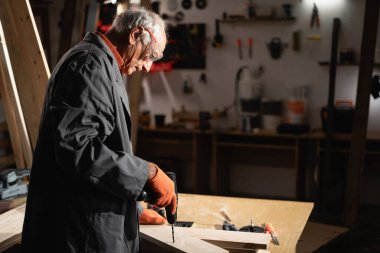 Güvenlik gözlüğü ve eldiven takan deneyimli marangoz bir atölyede ahşap kalaslar üzerinde çalışmak için elektrikli matkap kullanıyor. Sanatçılık, deneyim ve ahşap işçiliği kavramı