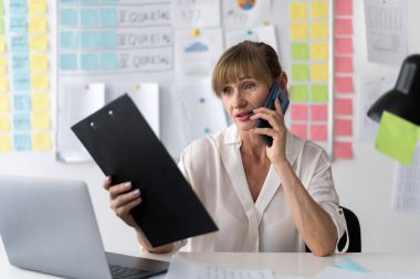 50 yaşındaki kurumsal bir kadın telefon görüşmesi yapıyor. Küçük ofisin içinde beyaz bir bluz giyiyor. Boşluğu kopyala