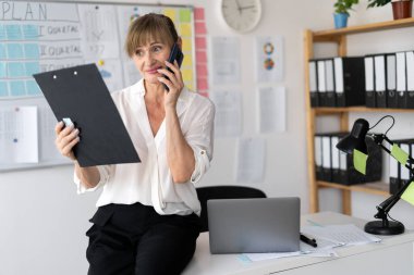 Bilgisayarlı kıdemli iş kadını ofiste çalışan bir grafiği analiz ediyor cep telefonu, iş ve mali konsept üzerinden arama yapıyor..
