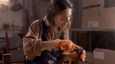 Bir atölyedeki kadın restorasyon projesinin bir parçası olarak verniğin kalıntılarını dikkatlice koltuktan kaldırıyor. O kahverengi bir gömlek, mavi önlük ve turuncu eldivenler giyer, işine odaklanır..