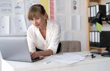 Ofiste çalışan yaşlı bir kadın dizüstü bilgisayarın üzerine eğilip bir rapora veri giriyor. Boşluğu kopyala