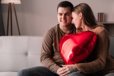 Sevgililer Günü kutlamanız kutlu olsun. Aşık genç bir çift evdeki kanepede otururken kalp şeklinde balon tutuyor. Boşluğu kopyala