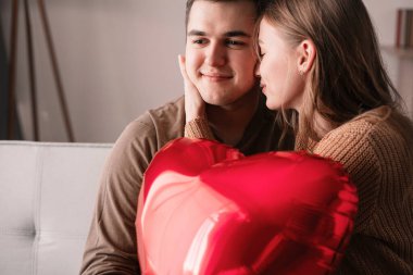 Sevgililer Günün kutlu olsun. Genç çift, oturma odasındaki kanepede oturan kalp şeklinde balona aşık. Evde romantik bir akşam. Boşluğu kopyala