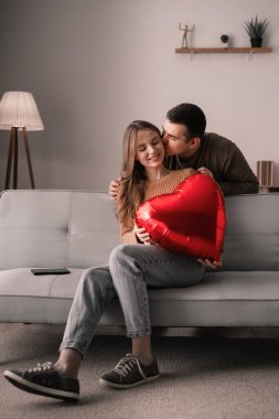 Sevgililer Günün kutlu olsun. Genç bir çift oturma odasındaki kanepede oturuyor, kız kırmızı kalp şeklinde balon tutuyor ve erkek kız arkadaşını öpüyor. Romantik bir akşam ve evde bir randevu.