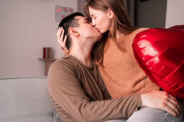 Aşık bir çift oturma odasında kanepede oturan kalp şeklinde balon tutarak öpüşüyor. Sevgililer Günü kutlaması konsepti