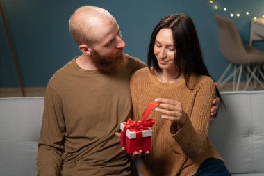 Sevgi dolu bir çift sıcak bir koltukta oturmuş kırmızı kurdeleye sarılmış özel bir hediyeyi değiş tokuş ediyorlar. Sevgiyi ve sıcaklığı yakalar. Rahat, samimi bir yuva ortamında..