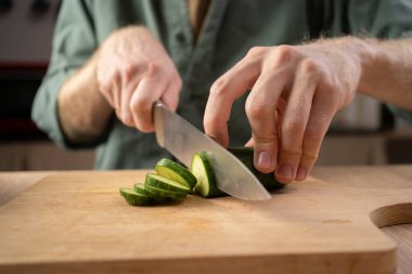 Keskin bir mutfak bıçağı ve tahta kesme tahtası kullanarak salataya salatalık kesmek için erkek eli yakın çekim. Boşluğu kopyala