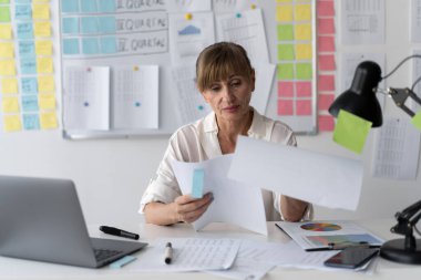 Ofis ortamında masasında çalışan yaşlı bir kadın. Arka planda beyaz tahtası var. O bir dizüstü bilgisayar kullanırken belgeleri tutuyor. Boşluğu kopyala