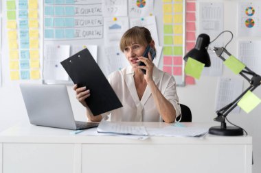 Ofiste çalışan yaşlı bir iş kadını dizüstü bilgisayar kullanıyor, cep telefonunu arıyor, emri kabul ediyor, belge doğrulamasını onaylıyor. Boşluğu kopyala