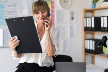 Profesyonel kıdemli iş kadını iş ortağıyla telefonda konuşuyor, ofisinde çalışıyor. Boşluğu kopyala