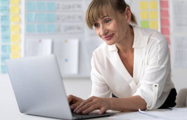 Ofiste duran dizüstü bilgisayarını kullanarak gülümseyen kıdemli iş kadını. Boşluğu kopyala