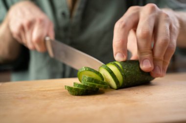 Sağlıklı beslenme ve beslenme konsepti. Erkek elleri evdeki mutfakta ahşap tahta üzerinde salatalık kesiyor, yakın plan.