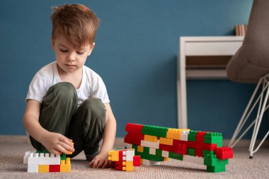 Mavi duvarları olan bir odada, yerde renkli plastik blokları olan odaklanmış küçük bir çocuk. Çocuk yaratıcı oyunlara, motor beceriler ve hayal gücü geliştirmeye derinden bağlıdır..