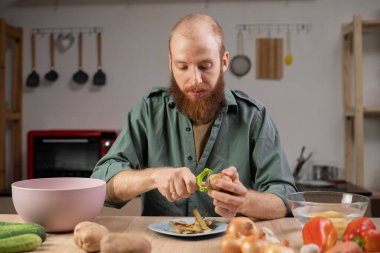 Sakallı adam ev mutfağında yemek yapıyor, patates ve diğer sebzeleri soyuyor, öğle yemeği için çorba hazırlıyor. Boşluğu kopyala