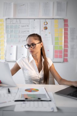 Masasındaki belgeleri inceleyen gözlüklü genç bir kadın, modern ofis ortamında çizelgeler ve grafiklerle çevrili, mesleki rolünde kendini adamış ve analitik becerilerini gösteriyor..