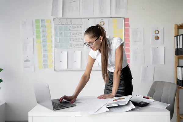 stock image Businesswoman in glasses working on a laptop, standing in a modern office with charts and plans on the wall. Represents dedication and attention to detail in a professional setting.