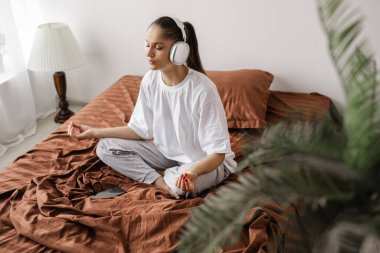 Yatakta kulaklıklı genç bir kadın müzik dinler meditasyon yapar, el ele tutuşur, dinlenir ve yatak odasında dinlenir. Yatakta uyan. Boşluğu kopyala