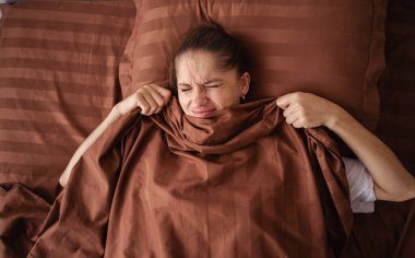 Young woman lies in bed, covering her face with a brown blanket. Her expression shows reluctance to wake up, capturing a relatable moment of wanting to stay cozy in bed on a chilly morning. clipart