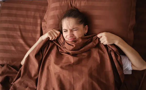 Stock image Young woman lies in bed, covering her face with a brown blanket. Her expression shows reluctance to wake up, capturing a relatable moment of wanting to stay cozy in bed on a chilly morning.