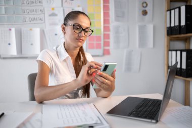 Ofiste dizüstü bilgisayarla çalışırken akıllı telefon kullanan bir iş kadını. İş yerinde çalışan bir kadın telefonunu kontrol ediyor. Kadın girişimci cep telefonuna göz gezdiriyor. Boşluğu kopyala