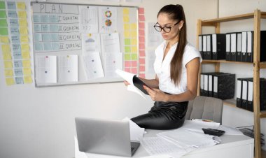 Gözlüklü bir muhasebeci ve beyaz bir bluz ofiste bir masanın üzerinde oturuyor ve bir panoda mali belgeleri dikkatle inceliyor. Haritalar, grafikler ve planlama malzemeleriyle çevrili.