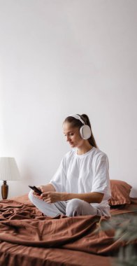 Genç bir kadın beyaz t-shirt ve kulaklık giyer yatakta oturup müzik dinler, yatak odasında akıllı telefon dinlenmesi kullanır. Dikey görünüm