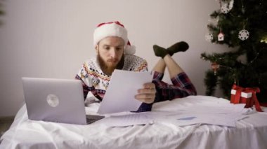 Noel Baba şapkalı bir iş adamı rahat yatağında oturuyor ve yılbaşı sabahı belgelerle dizüstü bilgisayarda çalışıyor. Boşluğu kopyala