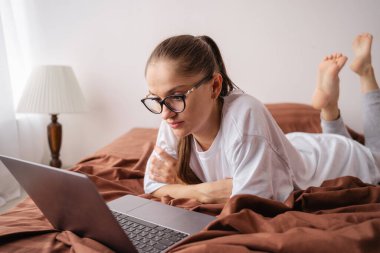 Gözlüklü genç bir kadın evde yatağında videolar ve sosyal medya içerikleri izlemek için dizüstü bilgisayar kullanıyor. Boş zaman, hafta sonu ve teknoloji konsepti