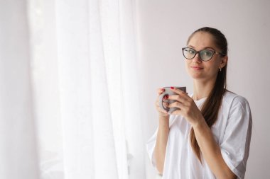 Bardaklı kadın pencerenin kenarında duruyor, elinde bir fincan çay ya da kahve tutuyor, sakin bir sabahın tadını çıkarıyor. Doğrudan kameraya bakıyor, sakin bir anı yakalıyor ve yansıtıyor..