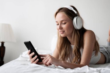 Rahatlatıcı teknoloji. Kablosuz kulaklıklı, yatak odasında dinlenirken ve cep telefonuyla müzik dinlerken akıllı telefon kullanan mutlu genç bir kadın. Boşluğu kopyala
