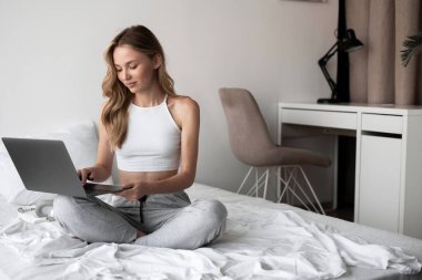 Beyaz bluzlu ve pijama pantolonlu genç bir kadın posta okuyor ve internette sohbet ediyor. Laptop bilgisayar kullanarak evde çalışıyor.