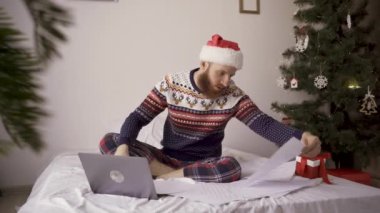 Noel süveteri ve Noel Baba şapkası giyen sakallı adam dizüstü bilgisayarda çalışıyor, belgelerle çalışıyor, şenlikli dekorasyonlarla çevrili, evdeki tatil ruhunu kucaklıyor.