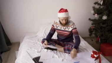İş adamı Noel Baba şapkası ve süveteri giyer. Belgeler ve dizüstü bilgisayarla çalışır. Yatakta oturur, evden çalışır.