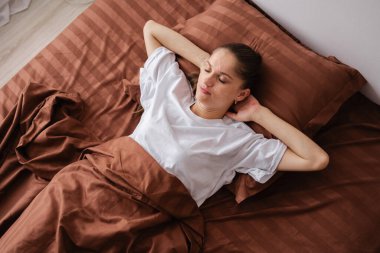 Esmer, uykucu bir kadın, güzel bir gece uykusundan sonra yatak odasında rahat bir sabahın keyfini çıkararak evde yatarak uyanıyor. Üst görünüm