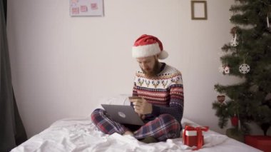 Noel Baba şapkalı genç adam yatak odasında oturmuş Noel alışverişi yaparken dizüstü bilgisayar kullanıyor. Çevrimiçi alışveriş ve tatil kavramı