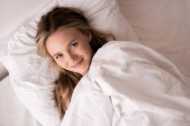 Beyaz battaniyenin altında uzanmış, gülümseyen ve iyi hisseden bir kadın. Boşluğu kopyala