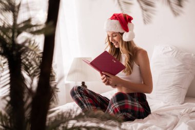 Noel Baba şapkası ve ekose pijamalı neşeli genç bir kadın yatağa oturmuş, pembe bir defter okuyor, rahat bir dekor ile çevrili, bayram sabahının tadını çıkarıyor.