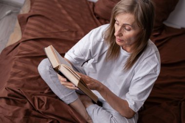 Son sınıf öğrencisi kadın beyaz t-shirt pijama giyer yatakta oturur ve kitap okur yatak odasında dinlenir. Boşluğu kopyala