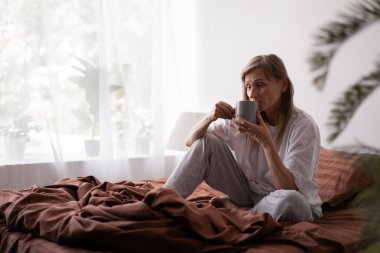 Evde, yatak odasında yatağında bir fincan kahveyle oturan yaşlı bir kadın. Günaydın, rahatlık ve insan konsepti.
