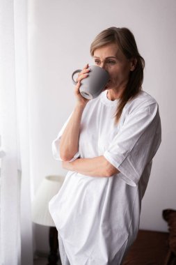 60 yaşında, rahat yatak odasında kahve içen yaşlı bir kadın hafta sonunun sakin ve huzurlu atmosferini kucaklıyor, rahatlık ve yalnızlığın tadını çıkarıyor.