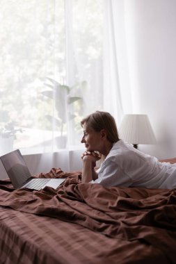 Yaşlı bir kadın dizüstü bilgisayar kullanarak yatağa uzanıyor. Yatak odasında bilgisayar kullanan mutlu bir kadın. Evde çalışmak ya da dizüstü bilgisayarla yatakta film izlemek. Boşluğu kopyala