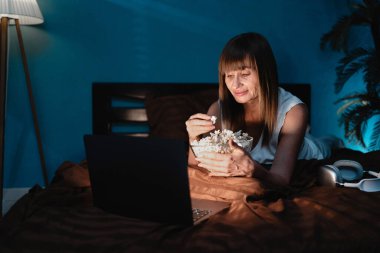 Geceleri yatakta orta yaşlı bir kadın dizüstü bilgisayarda televizyon izleyip patlamış mısır yiyor. Portre