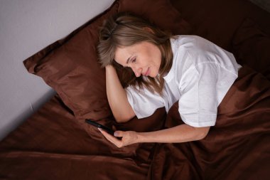 Yetişkin bir kadının telefonda yatakta dinlenmesi bunalımı. Yorgun, akıl hastası ve yalnız bir kadın yatak odasında sosyal medyada geziniyor. yüksek açı