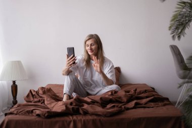 Son sınıf öğrencisi kadın akıllı telefonuyla yatakta oturuyor, ailesi ya da arkadaşlarıyla sohbet ederken mutlu bir şekilde el sallıyor, huzurlu ve rahat bir sabah geçiriyor..
