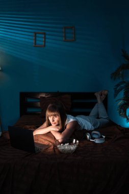 Orta yaşlı kadın patlamış mısır yiyor ve hafta sonu yatağında dinlenirken dizüstü bilgisayarda film izliyor. Boşluğu kopyala