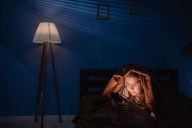 Sosyal medya için cep telefonu kullanan uykusuzluk çeken genç bir kız yatak odasındaki battaniyenin altında yatıyor. Boşluğu kopyala