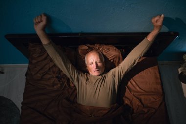 Yaşlı adam yatakta uyandıktan sonra kollarını açıyor, huzurlu bir yatak odasında dinleniyor, yeni güne hazırlanıyor, sabah ışığının keyfini çıkarıyor, rahat ve huzurlu uyandırma rutininin tadını çıkarıyor..