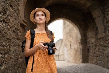 Kadın turist ve fotoğrafçı Avrupa 'daki yaz gezisi boyunca antik bir kalenin taş kemerinin yanında duruyor. Tarihsel güzellikleri yakalıyor, sıcak güneş ışığında eski yapıları keşfediyor..